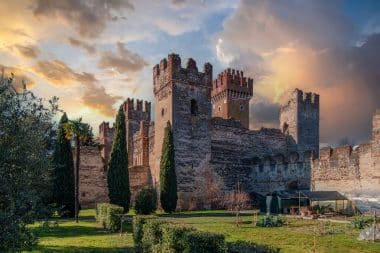 Eine alte Steinburg mit zinnenbewehrten Türmen und Mauern, umgeben von grünem Gras, hohen Zypressen und kleinen Gartenbauten vor dem Hintergrund eines dramatischen, farbenprächtigen Sonnenuntergangs.
