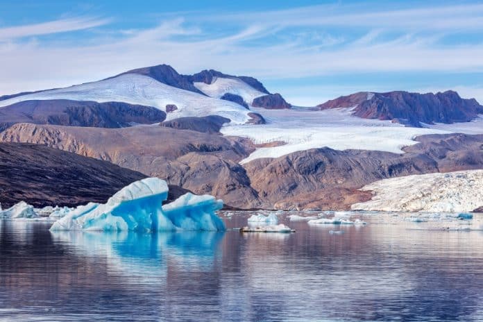 Svalbard,Landscape,Of,Glaciers,,Mountain,And,Blue,Icebergs,With,Reflections. Eisberge treiben in ruhigem Wasser, im Hintergrund sind ein Gletscher und felsige Berge zu sehen, über ihnen erstreckt sich ein blauer Himmel mit vereinzelten Wolken.
