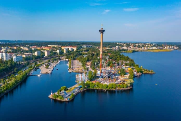Luftaufnahme eines Vergnügungsparks auf einer Halbinsel am Seeufer mit einem hohen Aussichtsturm – einer der bemerkenswerten Sehenswürdigkeiten von Tampere –, umgeben von Wasser, Bäumen und Stadtgebäuden unter einem strahlend blauen Himmel.