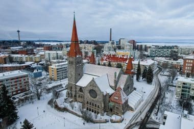 Eine große Steinkirche mit roten Dächern steht in einer verschneiten Stadtkulisse, umgeben von modernen Gebäuden und Bäumen. Schnee bedeckt den Boden und die Dächer, und die Szene strahlt eine kalte, winterliche Atmosphäre aus – ein perfektes Abbild einer der bekanntesten Sehenswürdigkeiten von Tampere.