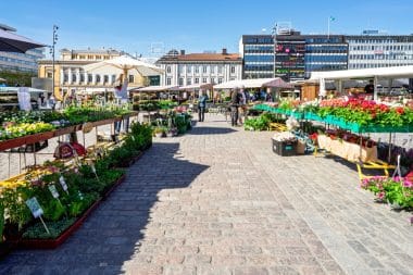 Ein Markt im Freien, auf dem verschiedene bunte Blumen und Pflanzen zum Verkauf angeboten werden, die auf Tischen entlang eines gepflasterten Gehwegs ausgestellt sind. Die Menschen stöbern unter Sonnenschirmen an den Ständen und genießen eine der reizvollen Sehenswürdigkeiten von Turku, während im Hintergrund an einem sonnigen Tag die Gebäude der Stadt zu sehen sind.