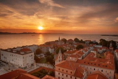 Luftaufnahme einer Küstenstadt mit roten Ziegeldächern bei Sonnenuntergang, mit Blick auf das ruhige Meer und die fernen Berge unter einem farbenprächtigen Himmel – eine der bezaubernden Sehenswürdigkeiten von Zadar, die die zeitlose Schönheit der Region einfängt.