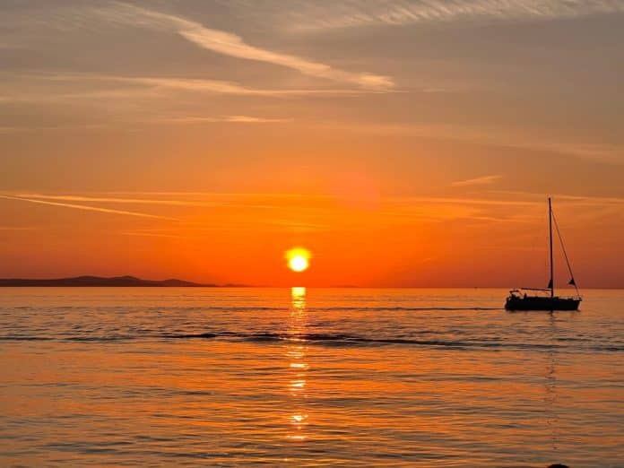 Watch,Sunset,In,Zadar,Croatia Ein Segelboot treibt auf ruhigem Wasser vor dem Hintergrund eines leuchtend orangefarbenen Sonnenuntergangs in der Nähe der Sehenswürdigkeiten von Zadar, während die Sonne tief am Horizont steht und sanfte Wolken den Himmel durchziehen.