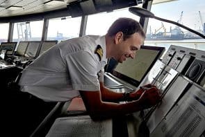 Ein Offizier in Uniform beugt sich auf der Brücke eines Schiffes über die Bedienfelder, überprüft Dokumente und elektronische Anzeigen, während die Fenster den Blick auf einen geschäftigen Industriehafen im Freien freigeben.
