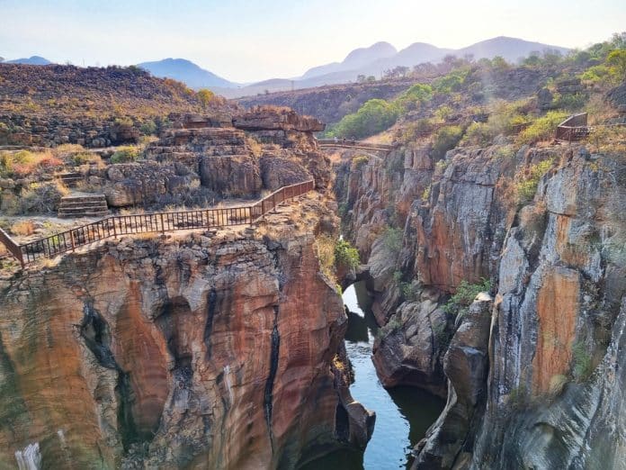 Eine felsige Schlucht mit tiefen, engen Schluchten, durch die ein Fluss fließt. Entlang der Klippen verlaufen mit Geländern gesicherte Stege, und im Hintergrund sind unter klarem Himmel Berge zu sehen.