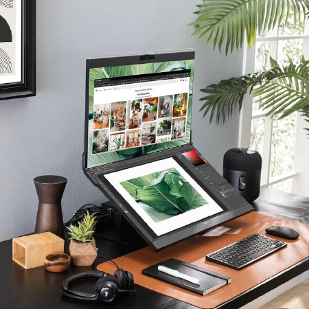 Eine moderne Schreibtischausstattung mit einem Monitor, einem Grafiktablett, auf dem ein Blattmotiv zu sehen ist, einer Tastatur, einer Maus, Kopfhörern, einem Smart-Lautsprecher und Schreibtischdekoration neben einem großen Fenster mit Sonnenlicht und grünen Pflanzen.