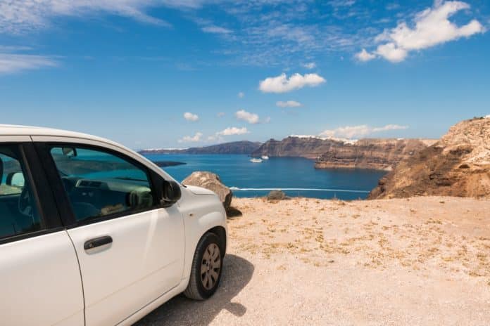 Santorini,Island,,Greece.,Panoramic,View,Of,The,Island,At,Sunny Ein weißes Auto, das auf einer Klippe geparkt ist, blickt auf das blaue Meer mit einem Boot und der fernen felsigen Küste unter einem sonnigen, teilweise bewölkten Himmel.