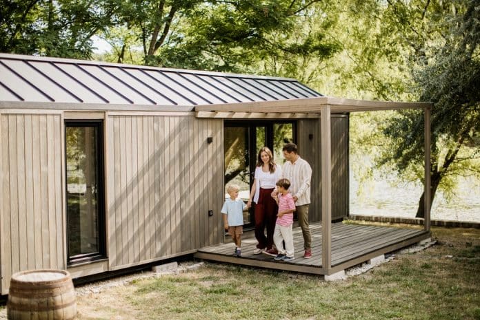 Eine vierköpfige Familie steht an einem sonnigen Tag auf der Veranda eines modernen Tiny House, das von Bäumen umgeben ist, lächelt und hält sich an den Händen.