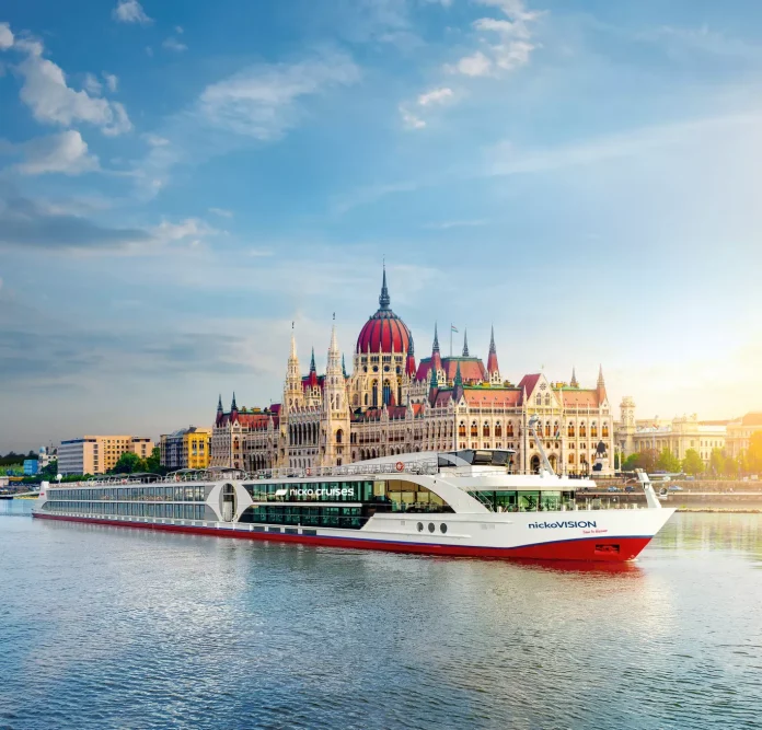 nickovision-budapest-parlament-sonnenaufgang-istock-1163188578-himmel-gefaerbt (1) Ein modernes Flusskreuzfahrtschiff gleitet auf einem ruhigen Fluss vor dem prunkvollen ungarischen Parlamentsgebäude in Budapest unter einem blauen Himmel mit leichten Wolken dahin.