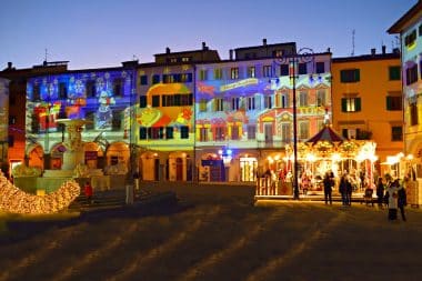 Ein belebter Stadtplatz in der Abenddämmerung mit farbenfrohen Weihnachtsprojektionen an den Gebäudefassaden, einem geschmückten Springbrunnen und einem hell erleuchteten Karussell, während die Menschen die festliche Atmosphäre genießen.