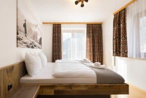 Ein Hotelzimmer mit einem Bett mit Holzrahmen, weißer Bettwäsche und gemusterten Vorhängen, die ein großes Fenster einrahmen.