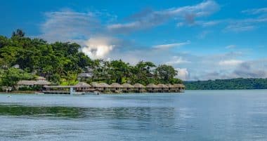 Eine Reihe von Überwasser-Bungalows entlang einer bewaldeten Küste unter blauem Himmel.