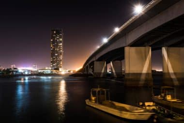 Nachtansicht einer Autobahnbrücke über ein ruhiges Gewässer mit Booten im Vordergrund und einem hohen Wohnhochhaus in der Ferne.