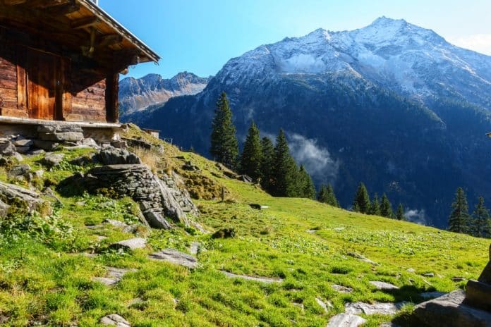 Eine rustikale Holzhütte auf einem grasbewachsenen Hang mit Blick auf schneebedeckte Berge.