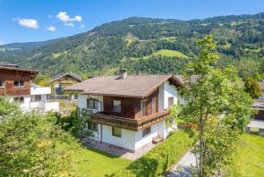 Ein zweistöckiges Haus mit einem Holzbalkon und einem braunen Ziegeldach, gelegen in einem Tal unterhalb grüner Berge.